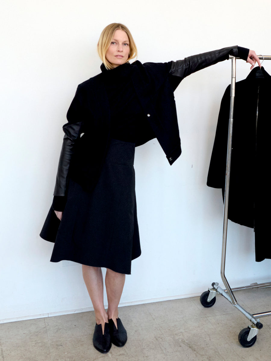 A woman stands indoors against a white wall, dressed in a black outfit featuring the Madi Skirt from Zero + Maria Cornejo, along with an asymmetrical hemline midi skirt, a top, and a jacket with leather sleeves. She poses with one arm raised and resting on a clothing rack that holds a black garment. The wooden floor beneath her adds warmth to the scene.