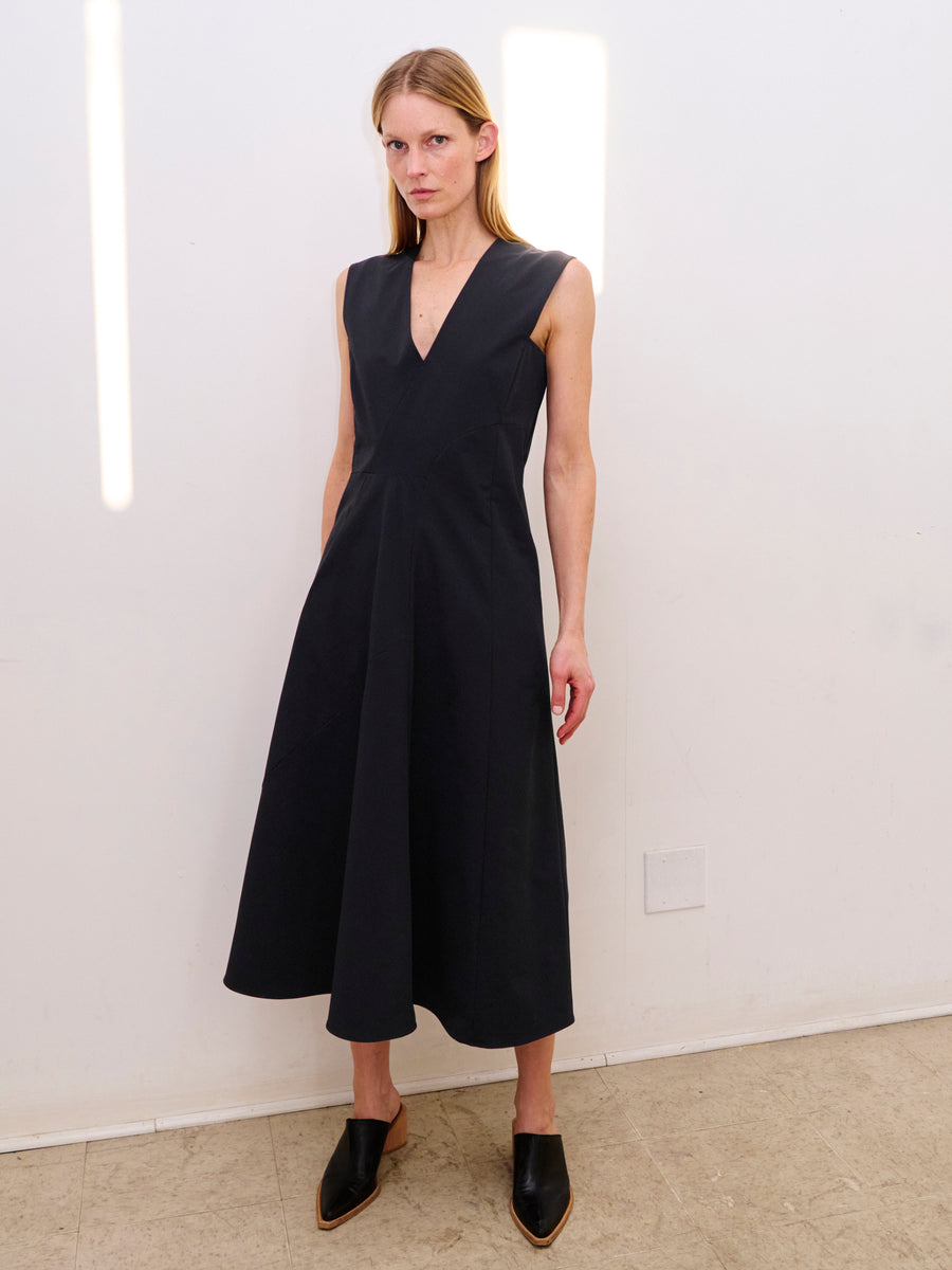 A person with long, light hair is standing against a white wall, wearing the sleeveless Eve Mosa Dress by Zero + Maria Cornejo crafted from technical stretch woven material and black slip-on shoes with a pointed toe. The person has a neutral expression and is standing on a tiled floor.