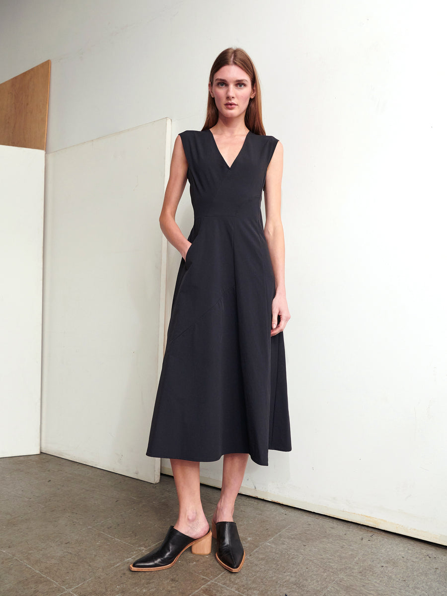 A person stands indoors against a plain, light-colored wall, wearing the Eve Mosa Dress by Zero + Maria Cornejo. The dress features a sleeveless V-neck design made from stretch woven fabric. They pair the dress with black heeled shoes and keep their hands casually tucked into the pockets, showcasing a minimalist aesthetic.