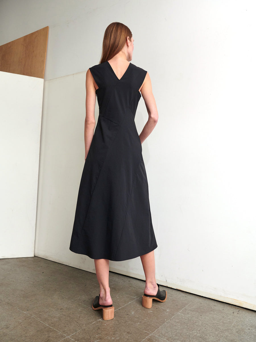 A woman wearing the Eve Mosa Dress by Zero + Maria Cornejo, made from stretch woven fabric, stands with her back to the camera in a minimalistic room. She pairs the sleeveless black dress with black slip-on heels and has long straight hair. The plain walls enhance the clean and simple aesthetic.