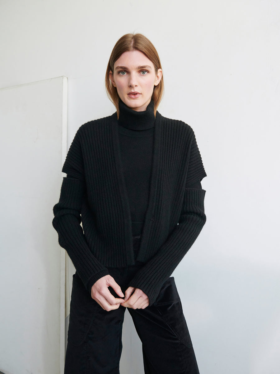 A person stands against a white wall, wearing the Zero + Maria Cornejo Slash Ani Cardigan over a black turtleneck and pants. They have straight, shoulder-length hair and are looking directly at the camera with a neutral expression.