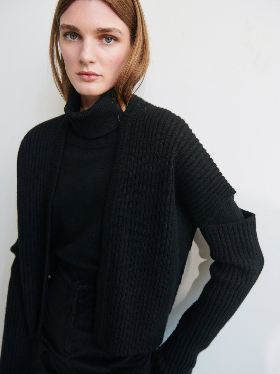 A person stands against a plain white background, wearing the Zero + Maria Cornejo Slash Ani Cardigan, a black ribbed knit piece featuring unique cutout details on the sleeves. They pair it with a black turtleneck underneath. The soft and lofty texture complements their shoulder-length light brown hair and neutral expression.