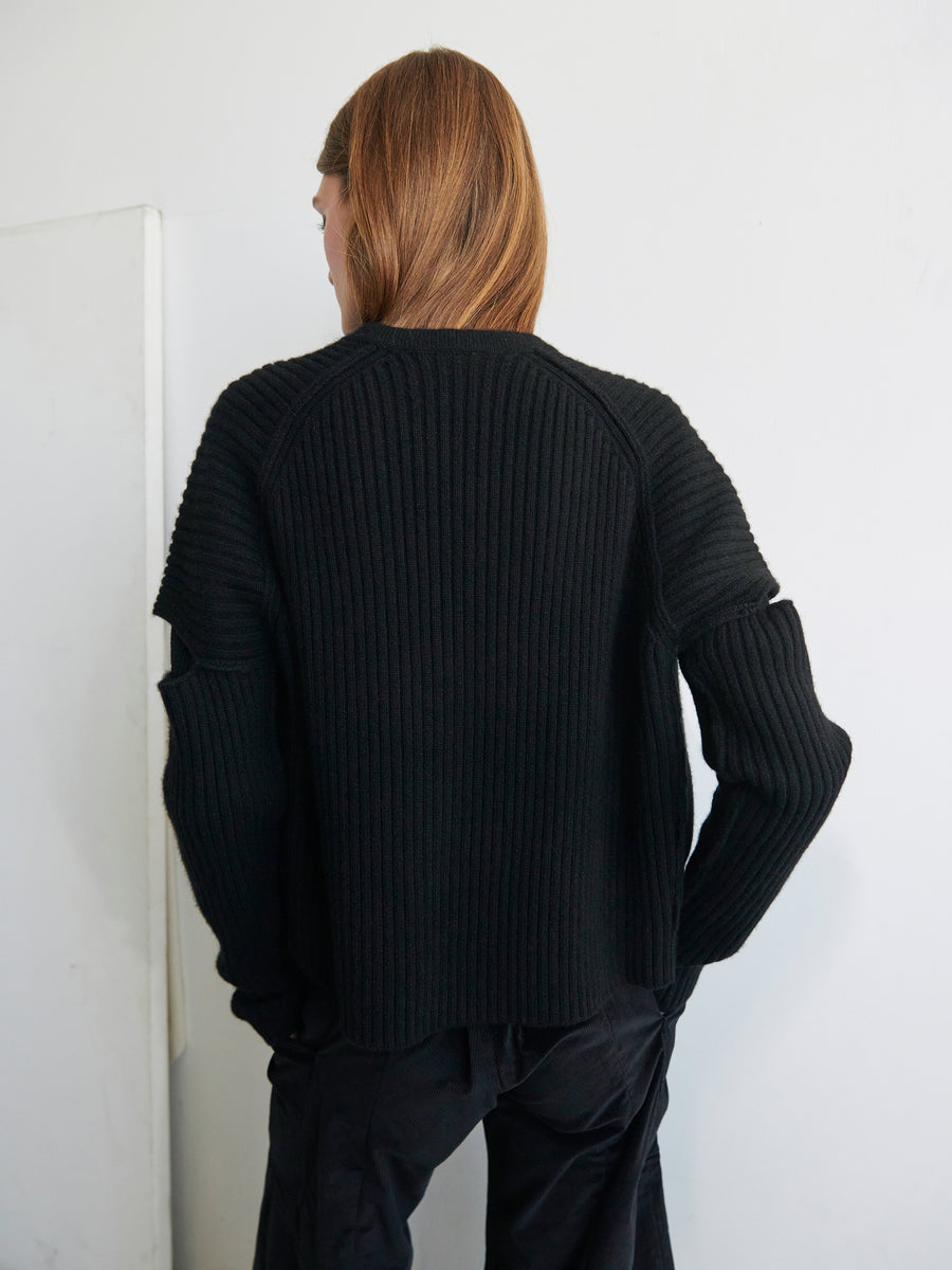 A person with long brown hair is standing facing away from the camera. They are wearing the Slash Ani Cardigan, a black ribbed knit cardigan made of recycled cashmere by Zero + Maria Cornejo, along with dark pants. The background is a plain, light-colored wall.