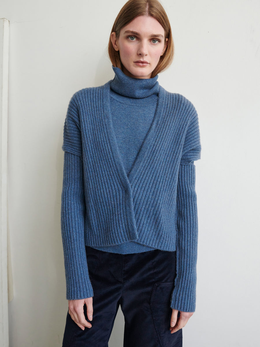 A person with shoulder-length hair is standing against a neutral background, wearing a blue recycled cashmere turtleneck sweater layered with the Slash Ani Cardigan from Zero + Maria Cornejo and dark pants. Their hands are relaxed at their sides, and they are looking into the camera.