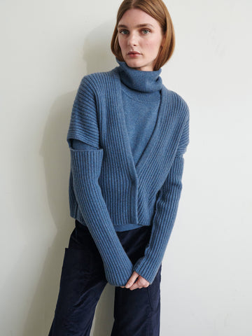 A person stands against a pale background in a blue Slash Ani Cardigan by Zero + Maria Cornejo and dark pants. Their shoulder-length hair partly obscures their face as they look away from the camera.