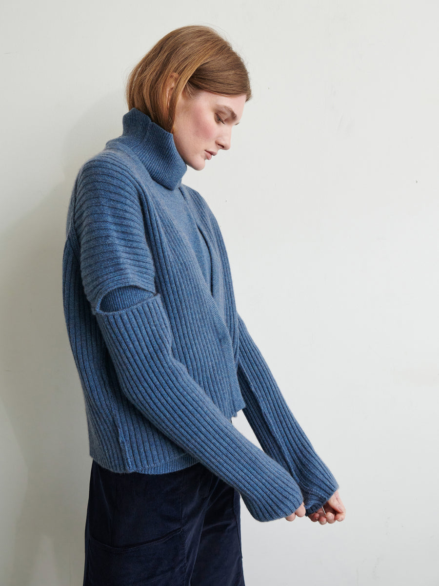 A person with short hair, wearing a blue turtleneck sweater and dark blue pants, stands against a plain light-colored wall with their head slightly turned to the side and looking down. One arm is bent while the other is slightly extended forward in a graceful manner, as if adjusting their Slash Ani Cardigan by Zero + Maria Cornejo.