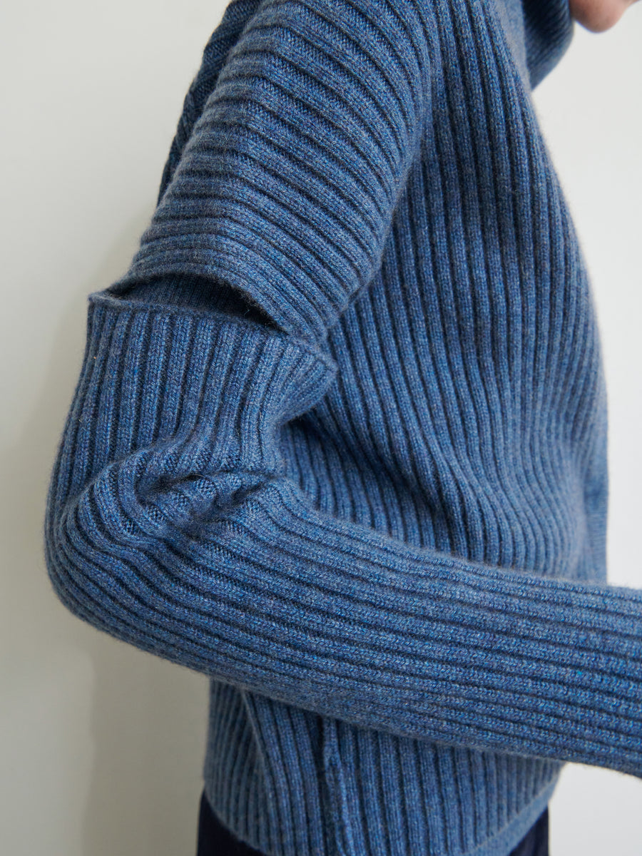 Person in a Slash Ani Cardigan by Zero + Maria Cornejo, showcasing its snug fit and vertical ribbing. This blue cardigan is made from recycled cashmere, with focus on texture and design against a plain light background.