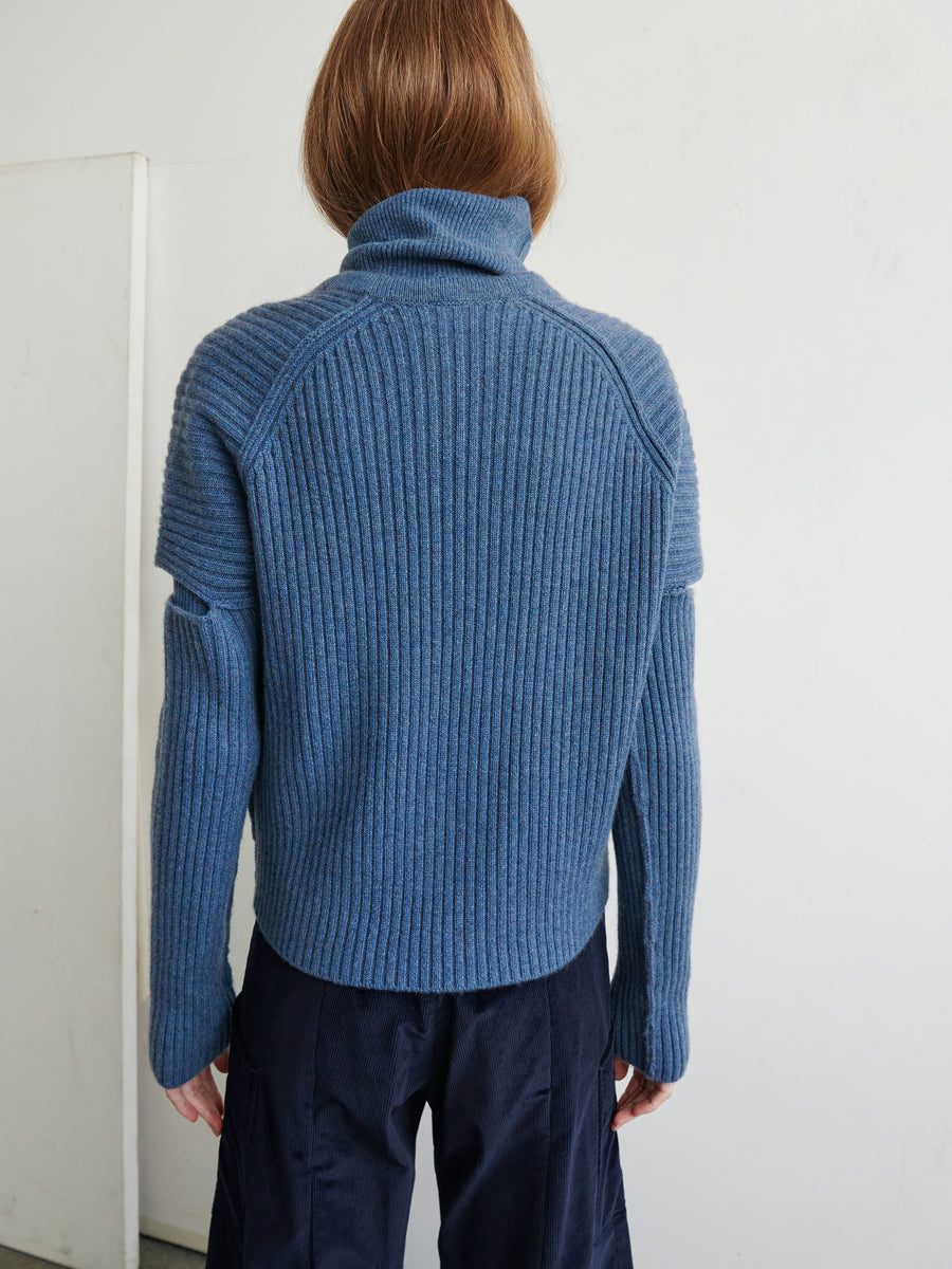 A person with shoulder-length hair is standing with their back to the camera, wearing a Slash Ani Cardigan by Zero + Maria Cornejo in blue and dark trousers. The setting appears to be indoors with a plain, light-colored wall.