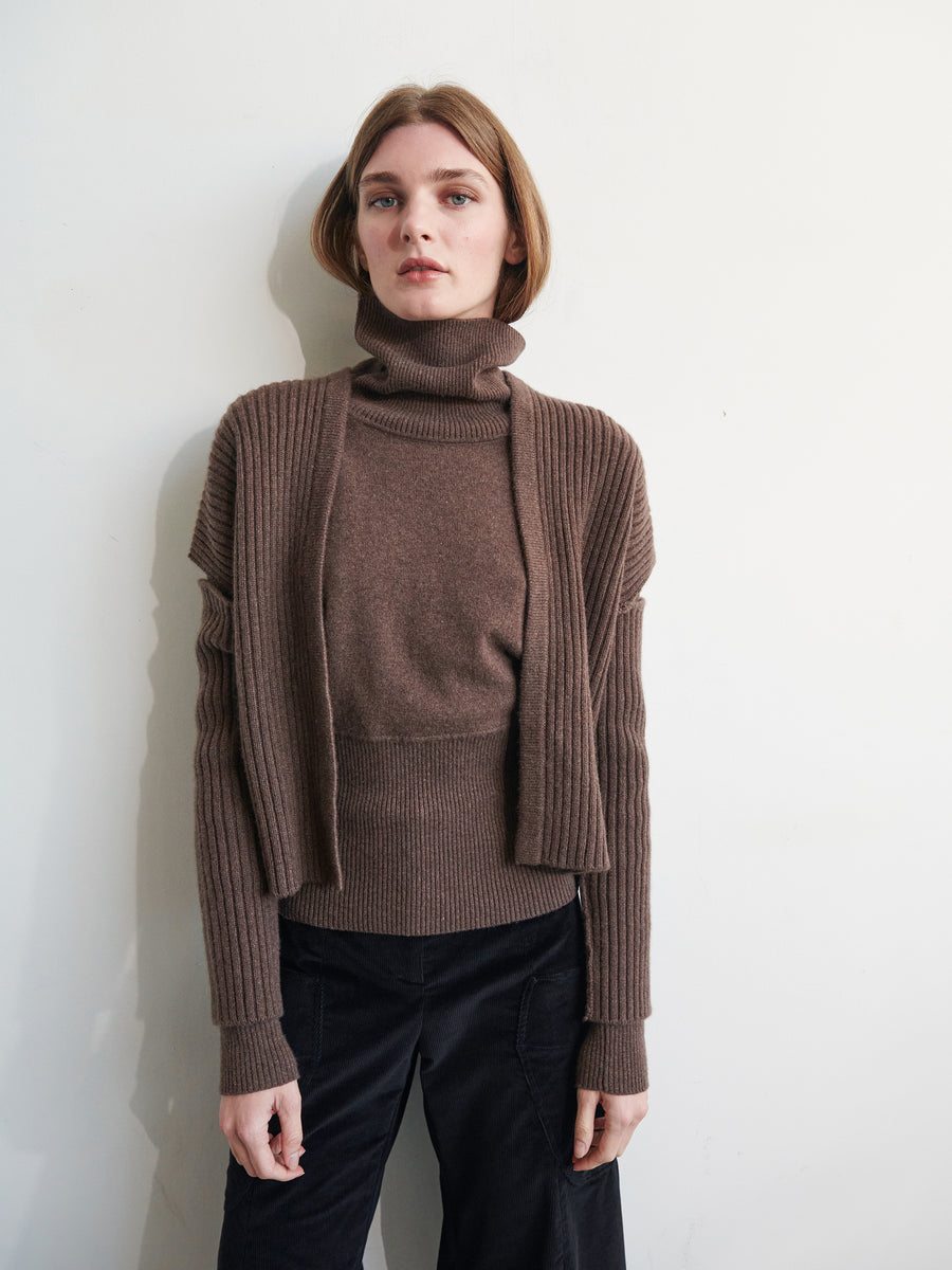 A person with short brown hair stands against a plain backdrop, wearing a brown recycled cashmere turtleneck sweater layered with the Slash Ani Cardigan by Zero + Maria Cornejo and black pants. They have a neutral expression, and their arms are relaxed at their sides.