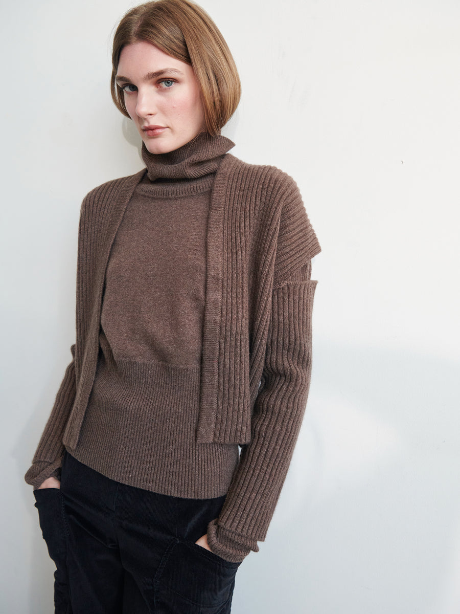 A person with short, light brown hair is wearing the Zero + Maria Cornejo Slash Ani Cardigan made from recycled cashmere and a matching ribbed turtleneck, paired with dark pants. They lean against a light wall, looking at the camera with hands in their pockets.