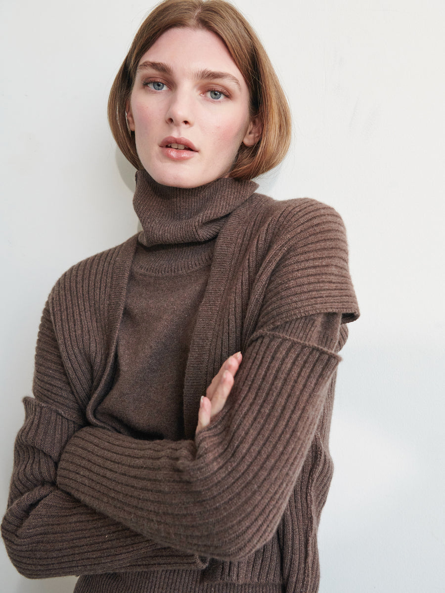 A person with short brown hair wears a brown ribbed turtleneck sweater and the Slash Ani Cardigan by Zero + Maria Cornejo. They stand against an off-white background, arms crossed, looking at the camera with a neutral expression.