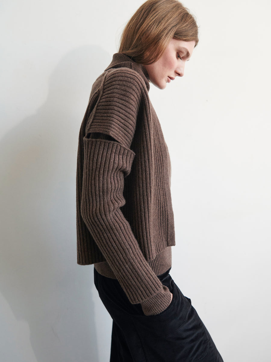 A person with long hair wears the Slash Ani Cardigan in brown by Zero + Maria Cornejo, paired with a black skirt, standing sideways with a hand in their pocket against a plain white background.