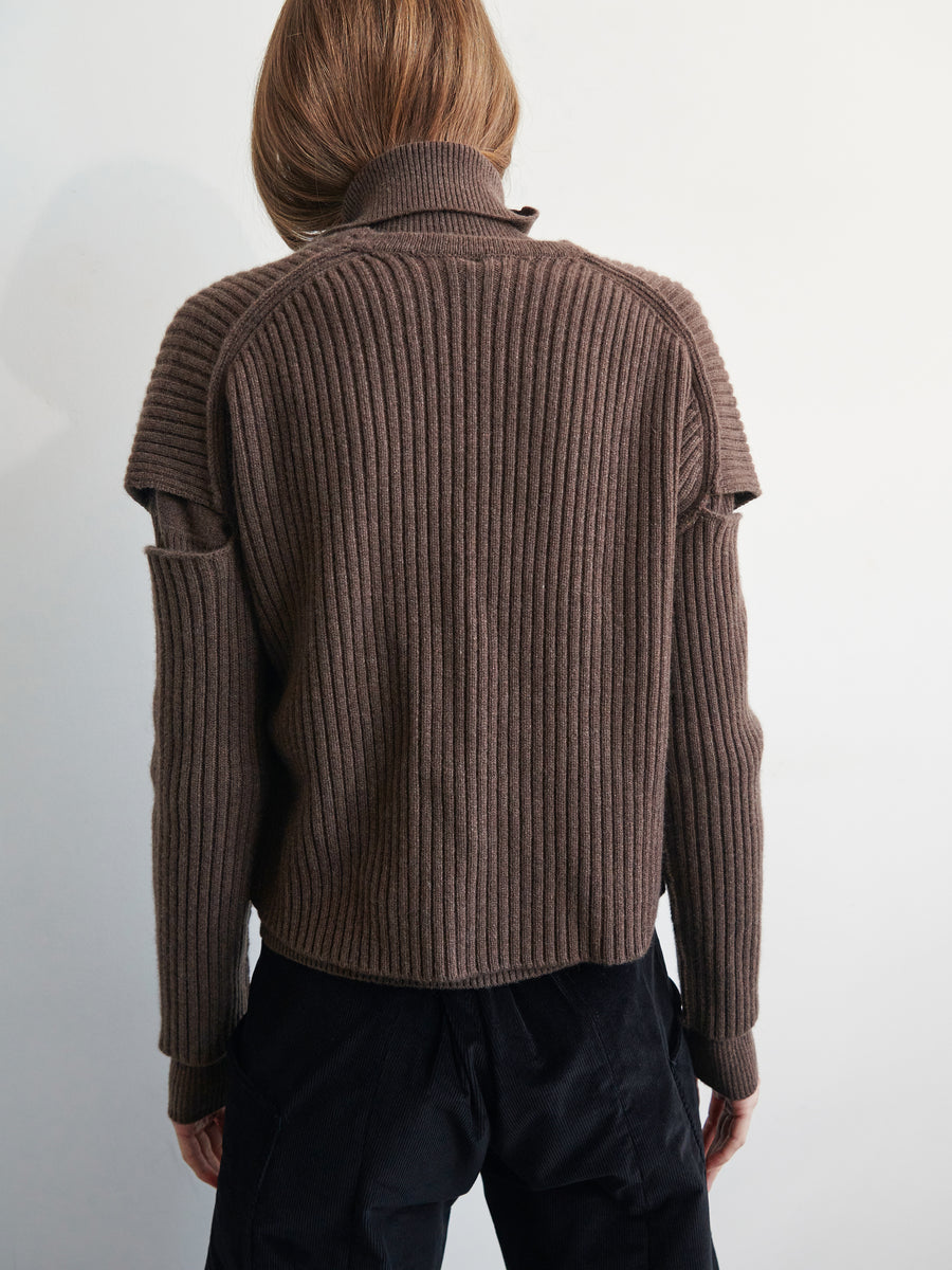 A person with long brown hair is seen from the back, wearing a Slash Ani Cardigan by Zero + Maria Cornejo, featuring distinctive shoulder cutouts and black pants. The soft and lofty texture of the cardigan contrasts beautifully against the plain, light-colored background.