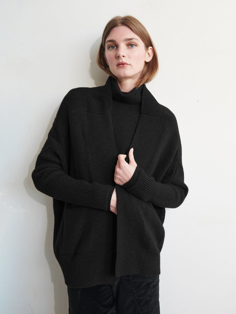 A person with short hair, against a plain light backdrop, wears a black turtleneck under the Orli Cardigan by Zero + Maria Cornejo. Their right hand keeps the cocoon silhouette closed while maintaining a neutral expression.