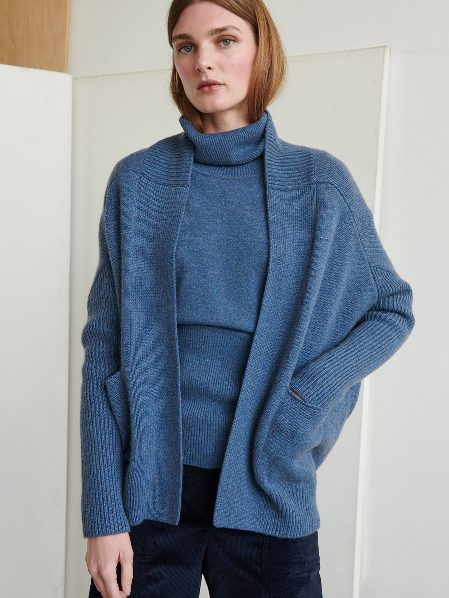 A person with shoulder-length light brown hair stands against a neutral background, wearing a cozy blue knit outfit. The outfit includes an ultra-soft cashmere turtleneck sweater paired with the Orli Cardigan by Zero + Maria Cornejo, featuring pockets and crafted from recycled cashmere, complemented by dark pants.