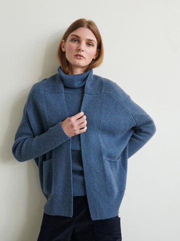 A person with shoulder-length brown hair is wearing a blue turtleneck sweater under the ultra-soft Orli Cardigan by Zero + Maria Cornejo. They are standing against a light-colored wall, looking directly at the camera with a relaxed expression.