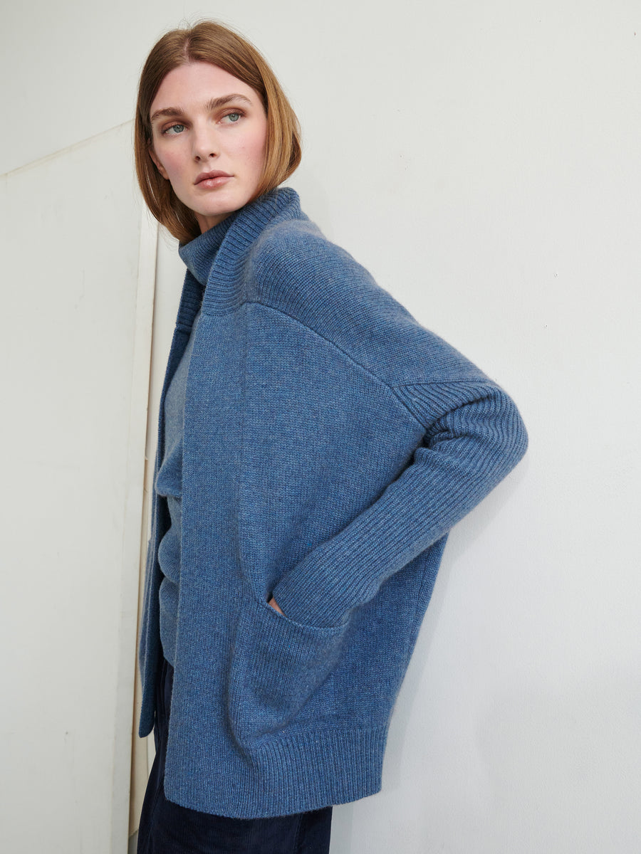 A person with shoulder-length hair is standing against a plain wall, wearing the ultra-soft Orli Cardigan from Zero + Maria Cornejo and a matching blue top. The recycled cashmere Orli Cardigan is open-front with pockets, and the person is looking slightly to the side with a neutral expression.