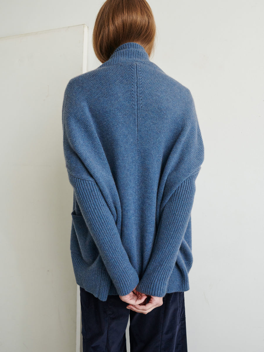 A person with long brown hair stands facing a white wall, wearing a loose, oversized blue Orli Cardigan from Zero + Maria Cornejo with their hands clasped behind their back. The ultra-soft cashmere cardigan has a high collar, and the person is dressed casually in dark pants.