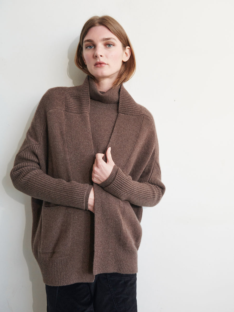 A person with shoulder-length hair is standing against a plain off-white wall. They are wearing an ultra-soft Orli Cardigan, from Zero + Maria Cornejo, in brown. The oversized open-front cardigan is draped over a matching brown turtleneck, with one hand holding the cardigan closed and the other hand partially visible.