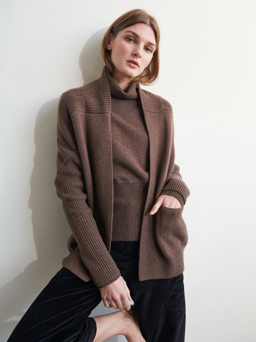 A person with short, light brown hair stands against a white wall. They are wearing the Orli Cardigan by Zero + Maria Cornejo, a brown, long-sleeved, knitted sweater with a cocoon silhouette over a matching turtleneck and dark pants. One hand is in their cardigan pocket as they gaze towards the camera.