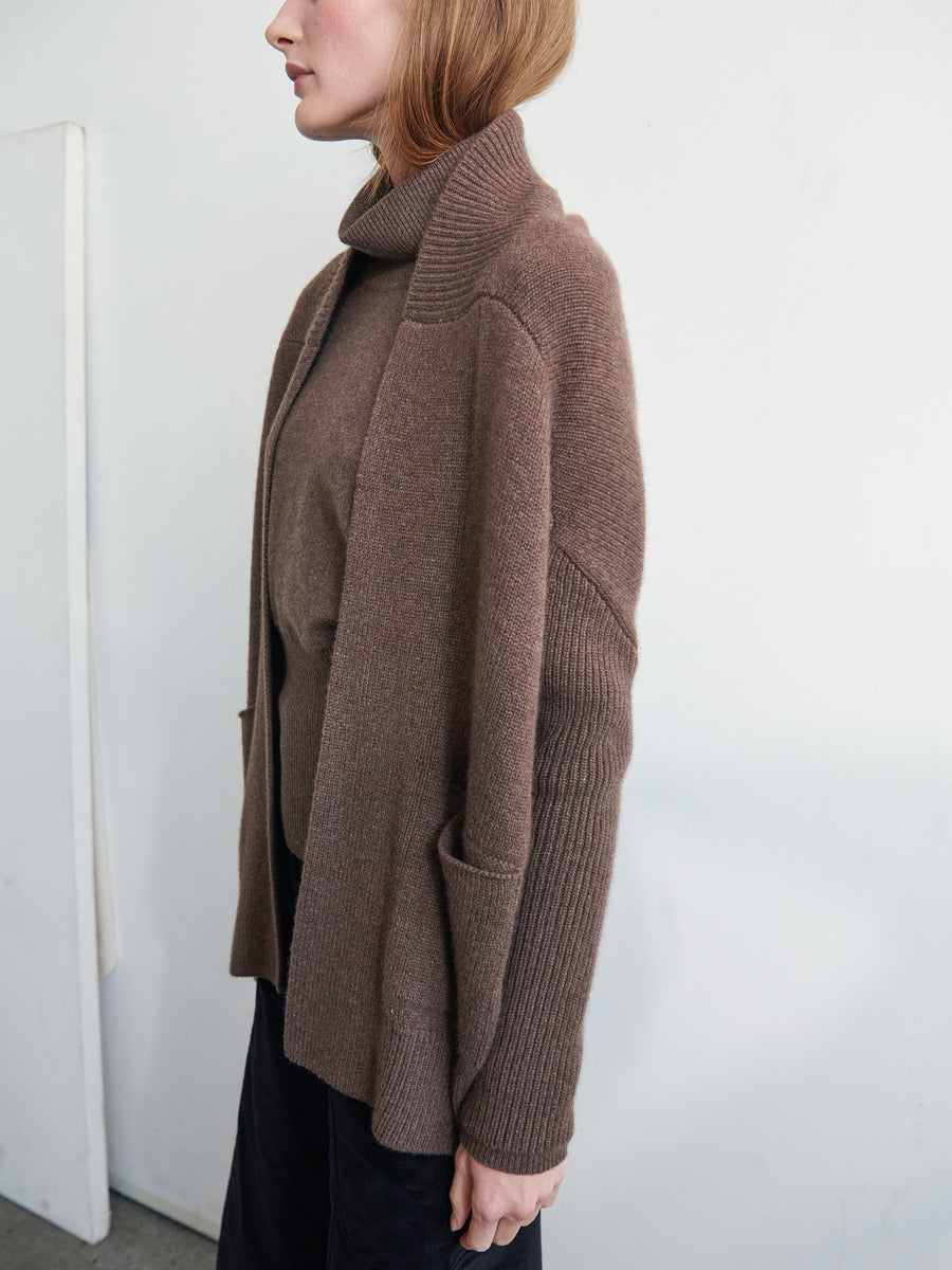 A person with shoulder-length light brown hair is wearing an ultra-soft cashmere turtleneck sweater and the Orli Cardigan by Zero + Maria Cornejo, featuring a cozy cocoon silhouette. They have their side to the camera, standing against a plain white background.