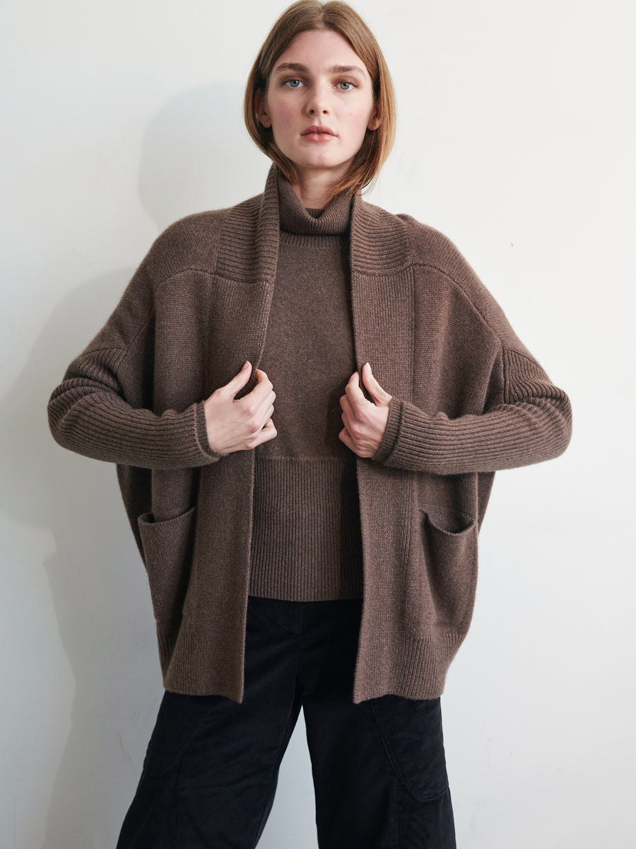 Standing against a plain backdrop, a person with shoulder-length hair models the ultra-soft Orli Cardigan by Zero + Maria Cornejo over a matching brown sweater. Their hands rest inside the pockets of the cocoon silhouette cardigan, and they are wearing black pants.