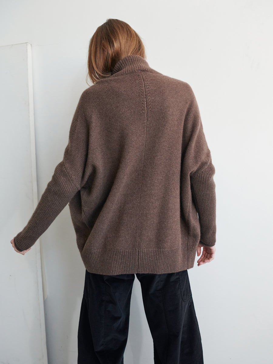 A person with long hair is seen from the back, wearing a loose Orli Cardigan by Zero + Maria Cornejo and black pants. They are positioned against a plain, white background. Their left arm is slightly extended.