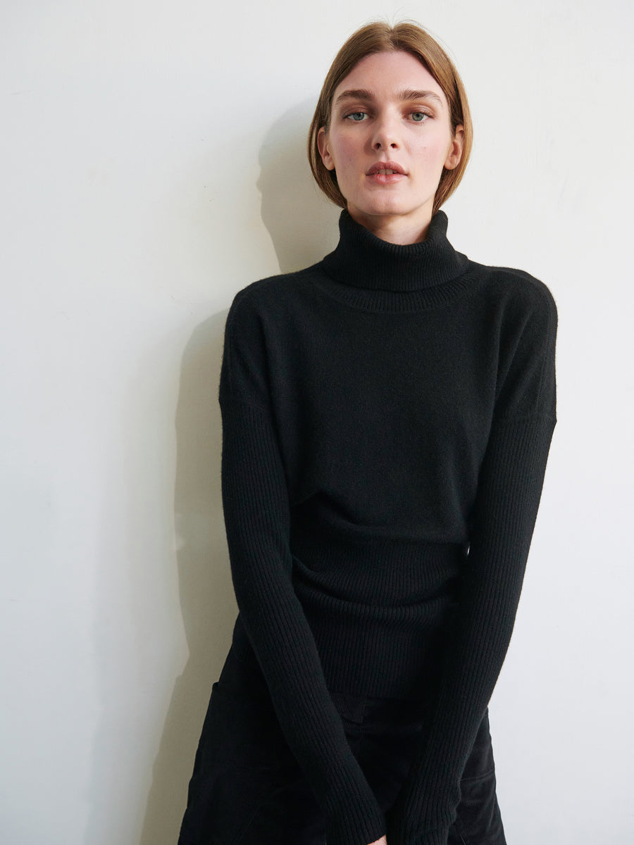 A person with shoulder-length hair is standing against a white wall, wearing a black Long-Sleeved Ama Rollneck by Zero + Maria Cornejo. Their arms are relaxed at their sides as they look directly at the camera. The lighting is soft, casting minimal shadows on the wall, highlighting their commitment to sustainable fashion.