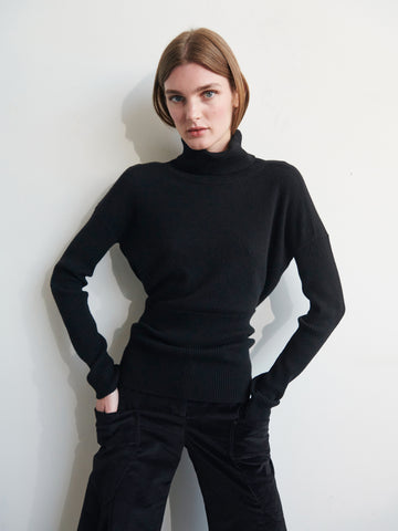 A person with short brown hair is wearing a black Long-Sleeved Ama Rollneck by Zero + Maria Cornejo and black pants. They are standing against a plain, light-colored background with their hands on their hips, looking directly at the camera with a serious expression.