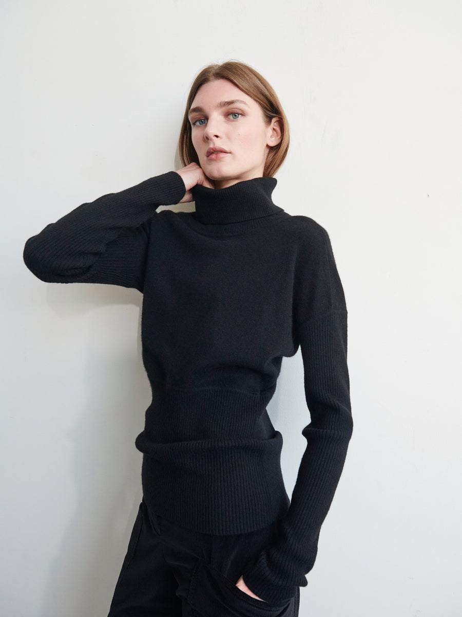 A person with straight, shoulder-length brown hair is standing against a plain wall, wearing the Long-Sleeved Ama Rollneck from Zero + Maria Cornejo made of recycled cashmere. They have one hand raised, touching their hair and are gazing forward with a neutral expression.