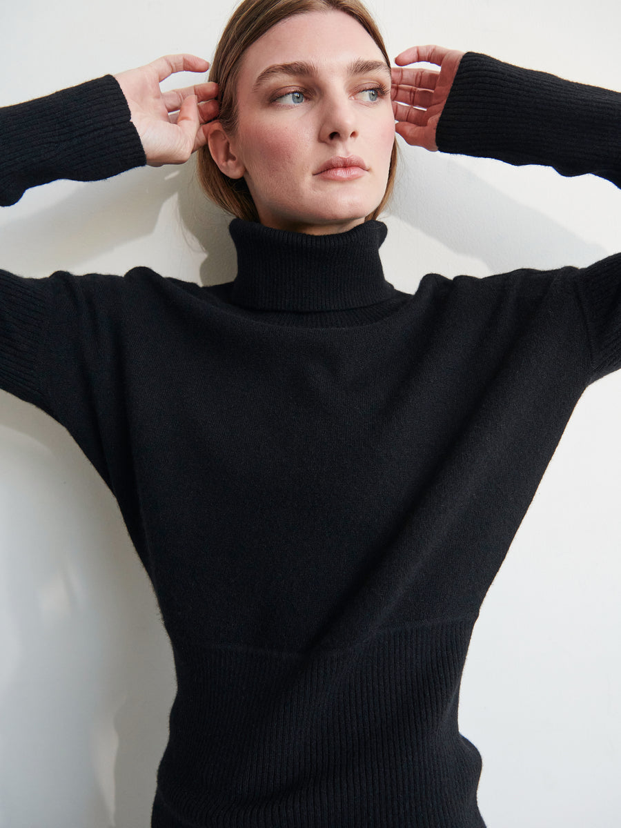 A person wearing the Long-Sleeved Ama Rollneck by Zero + Maria Cornejo, a black knit pullover with ribbed sleeves, is standing against a plain white background. They have light-colored hair pulled back, and are raising their hands near their head while looking slightly to the side.