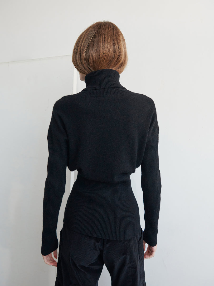 A person with short brown hair is standing facing away. They are wearing the fitted Long-Sleeved Ama Rollneck in black and matching black pants by Zero + Maria Cornejo. The background is plain white, highlighting the simplicity and elegance of sustainable fashion.