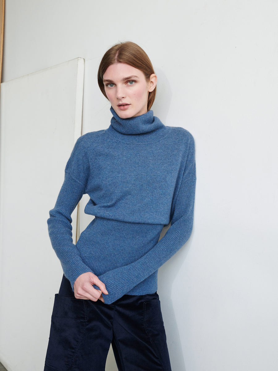 A person with straight, shoulder-length hair is standing against a plain wall. They are wearing the Long-Sleeved Ama Rollneck by Zero + Maria Cornejo, made of recycled cashmere, along with navy blue pants. They look directly at the camera with a neutral expression, their hands slightly clasped in front of them.