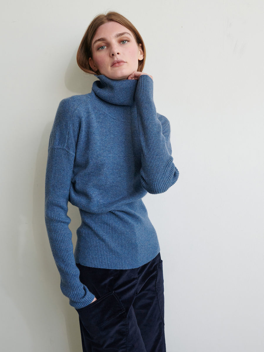 A person with short hair is wearing a Zero + Maria Cornejo Long-Sleeved Ama Rollneck in blue, crafted from recycled cashmere, along with dark pants while posing against a plain light-colored wall. One hand is partially inside the ribbed sleeve, and the other is relaxed by their side. They have a calm, contemplative expression.