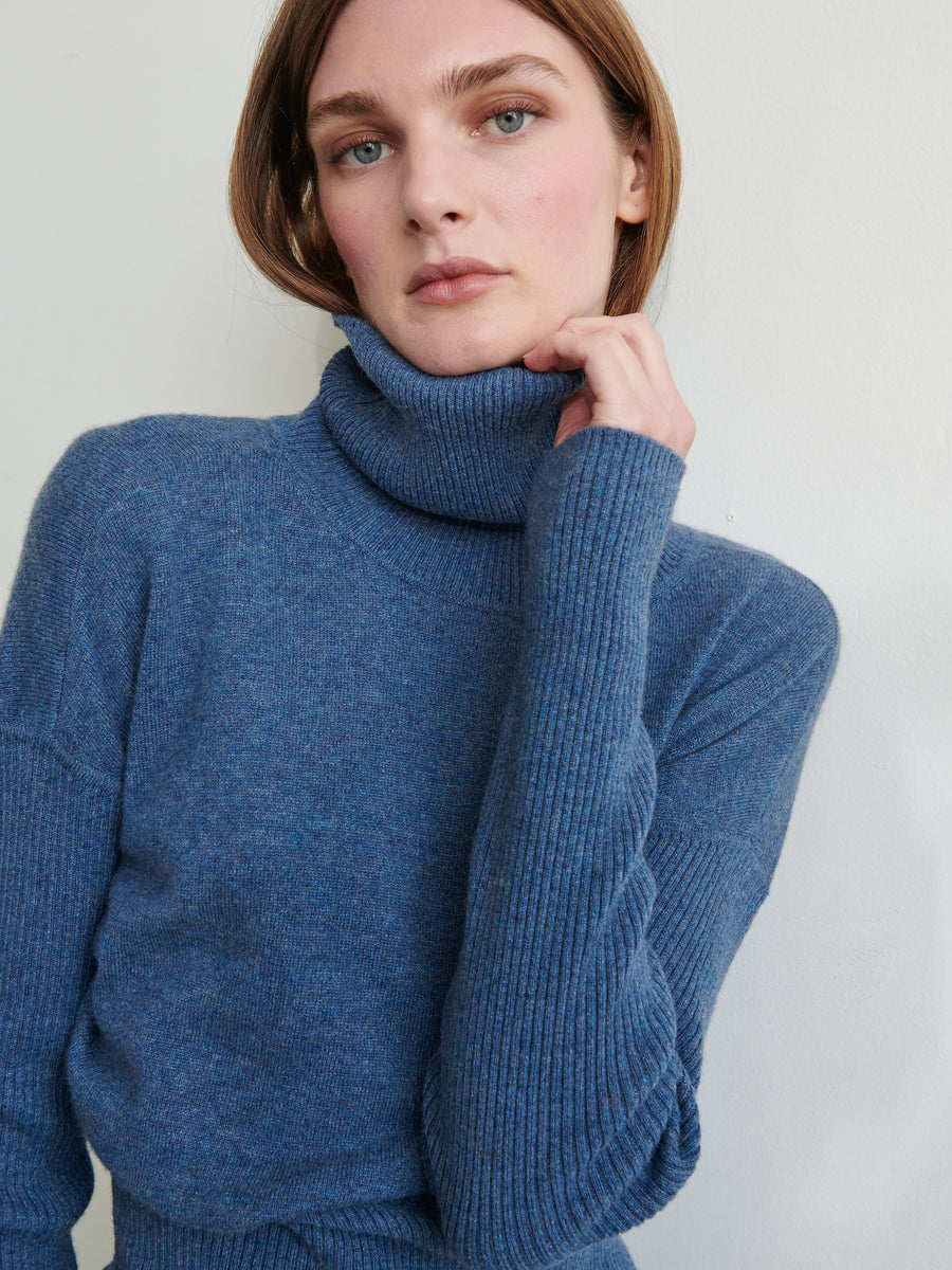 A person with short brown hair, dressed in a blue Long-Sleeved Ama Rollneck sweater from Zero + Maria Cornejo, gazes forward with a calm expression. One hand gently touches the cozy rollneck pullover made from recycled cashmere, while the background is a plain, light-colored wall.