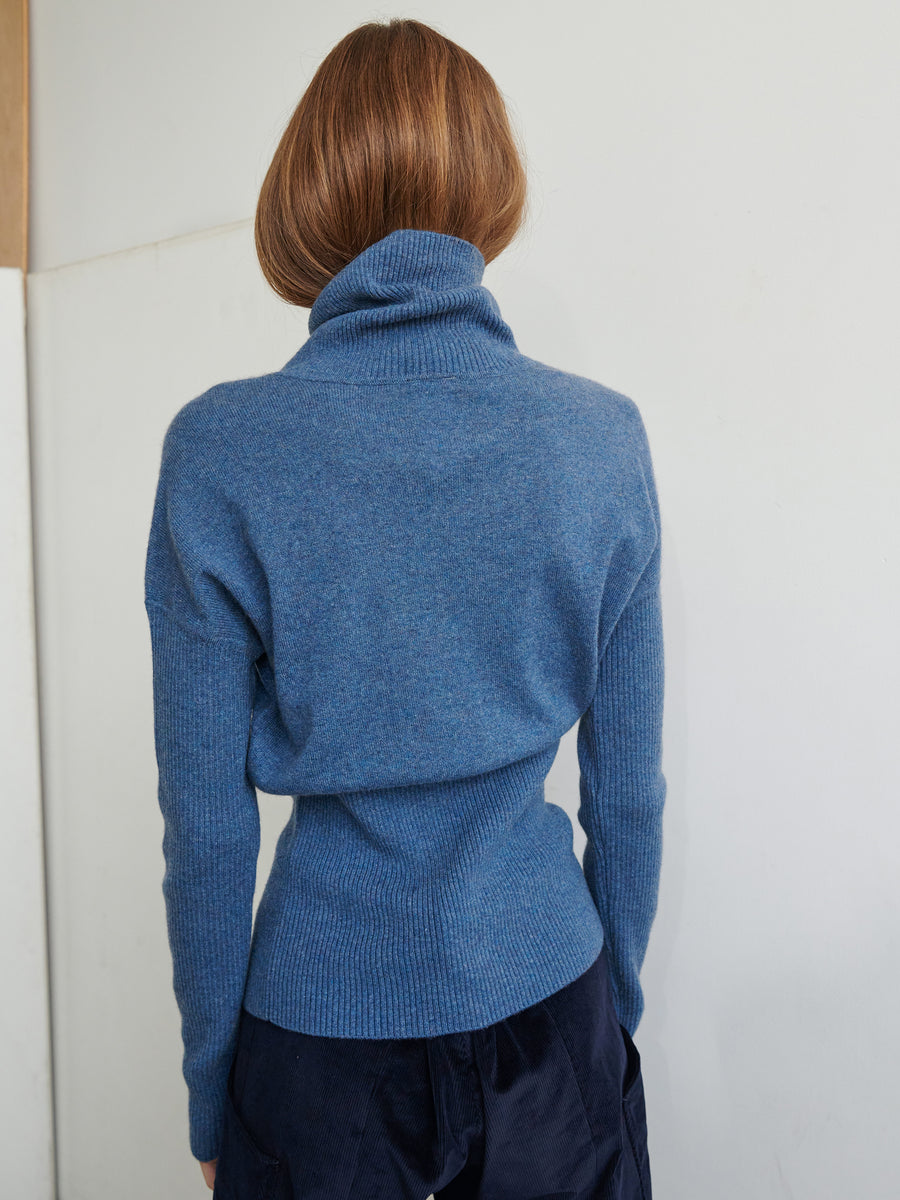 A person with shoulder-length brown hair is standing with their back to the camera, donning the Long-Sleeved Ama Rollneck by Zero + Maria Cornejo in blue, crafted from recycled cashmere paired with dark pants. The light-colored wall in the background accentuates the elegance of sustainable fashion.