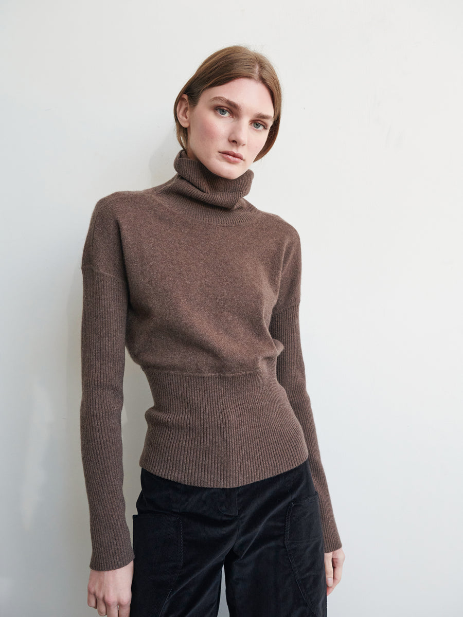 A person with brown hair is wearing a fitted Long-Sleeved Ama Rollneck by Zero + Maria Cornejo, paired with black pants. They are looking slightly to the side while standing against a plain, light-colored background.