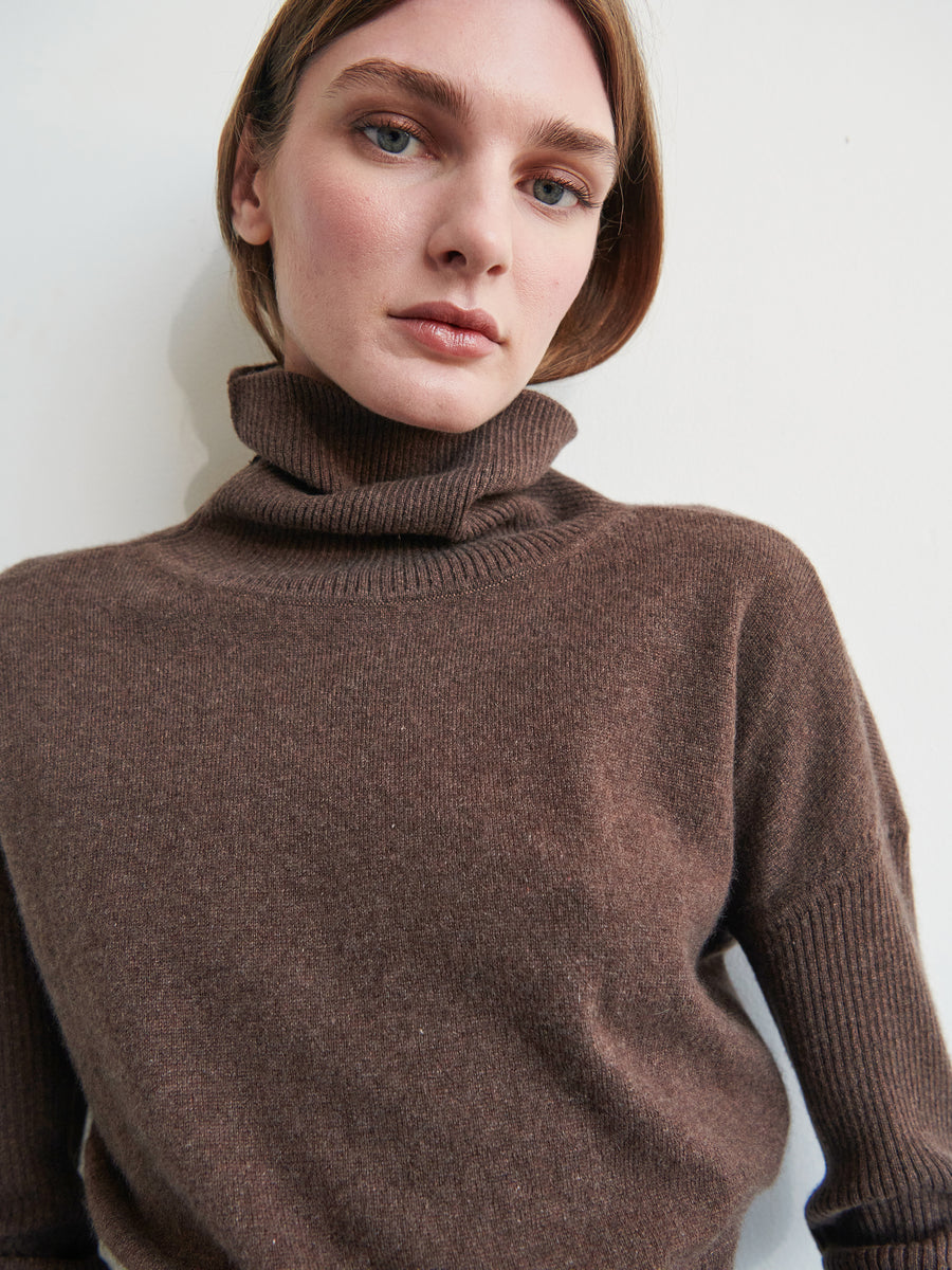 A person with fair skin and light brown hair is wearing the Long-Sleeved Ama Rollneck by Zero + Maria Cornejo. The ribbed sleeves add texture to the brown turtleneck sweater as they gaze directly at the camera against a plain, light background.