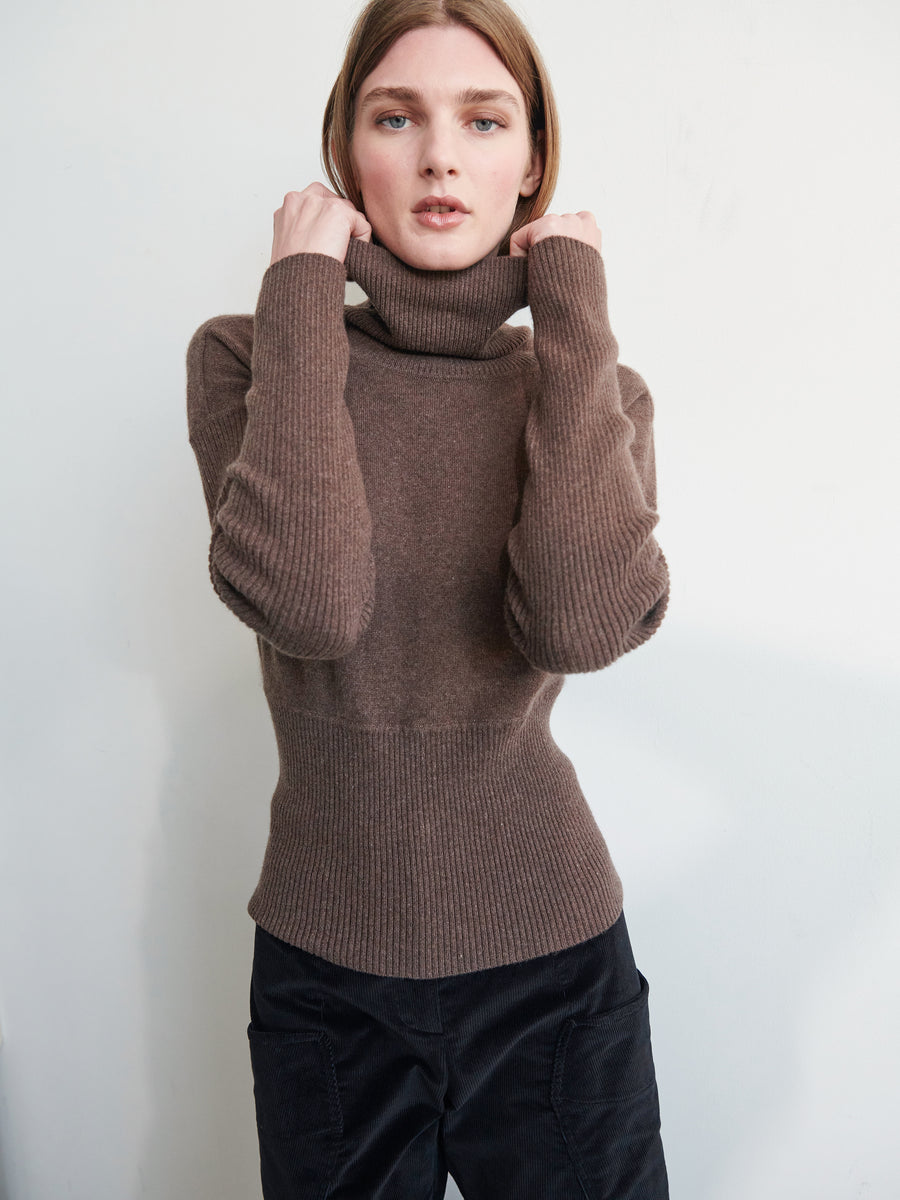 A person with shoulder-length hair poses in the Long-Sleeved Ama Rollneck by Zero + Maria Cornejo, featuring ribbed sleeves and hem. They have one arm bent upward to touch the raised collar while looking directly at the camera against a plain, light background.