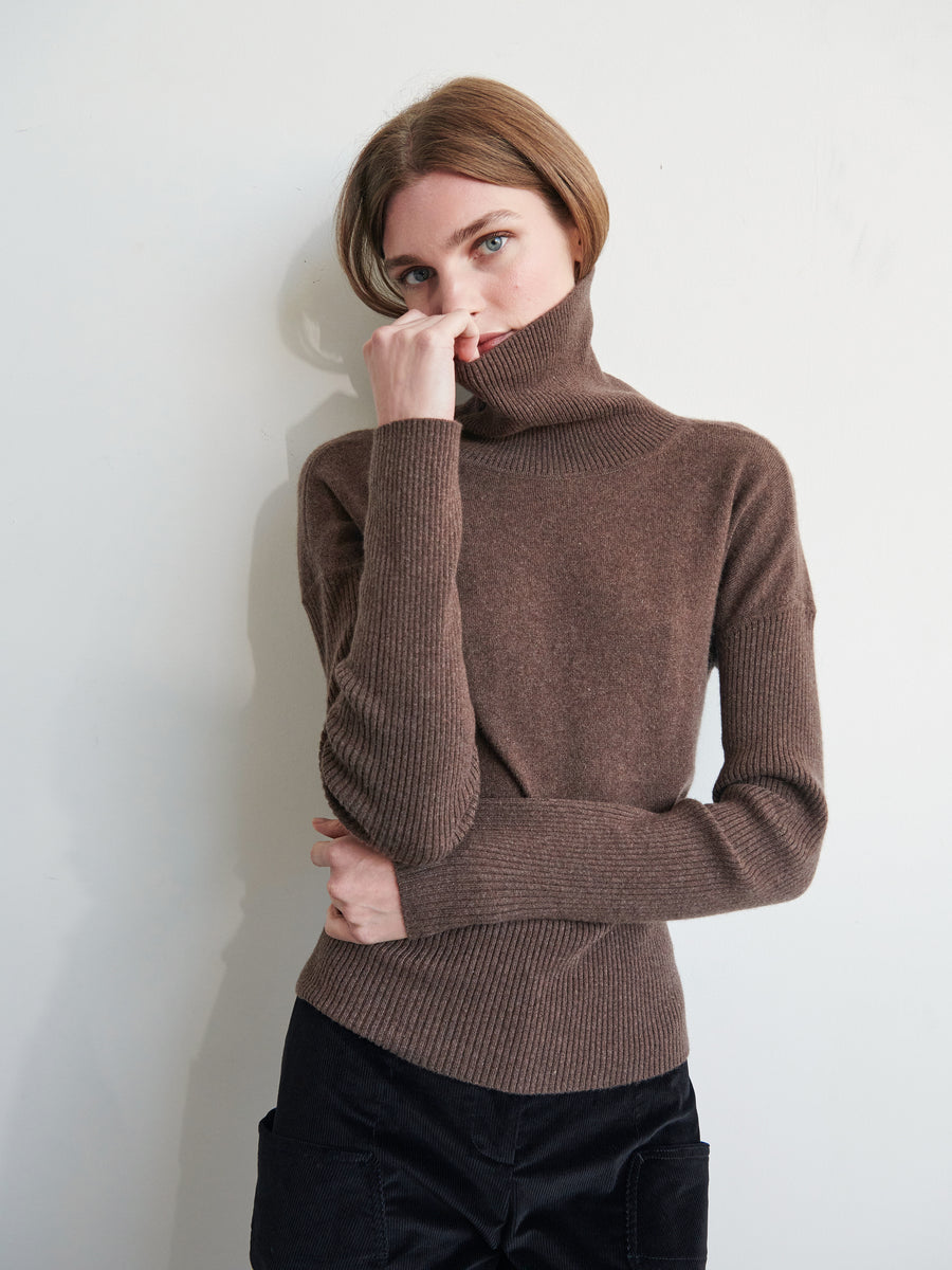 A person with light brown hair is wearing a Long-Sleeved Ama Rollneck pullover from Zero + Maria Cornejo, pulling the neck part up to their face. They have one hand covering their mouth and the other loosely folded across their waist. The ribbed sleeves add texture as they stand against a plain, light-colored wall.