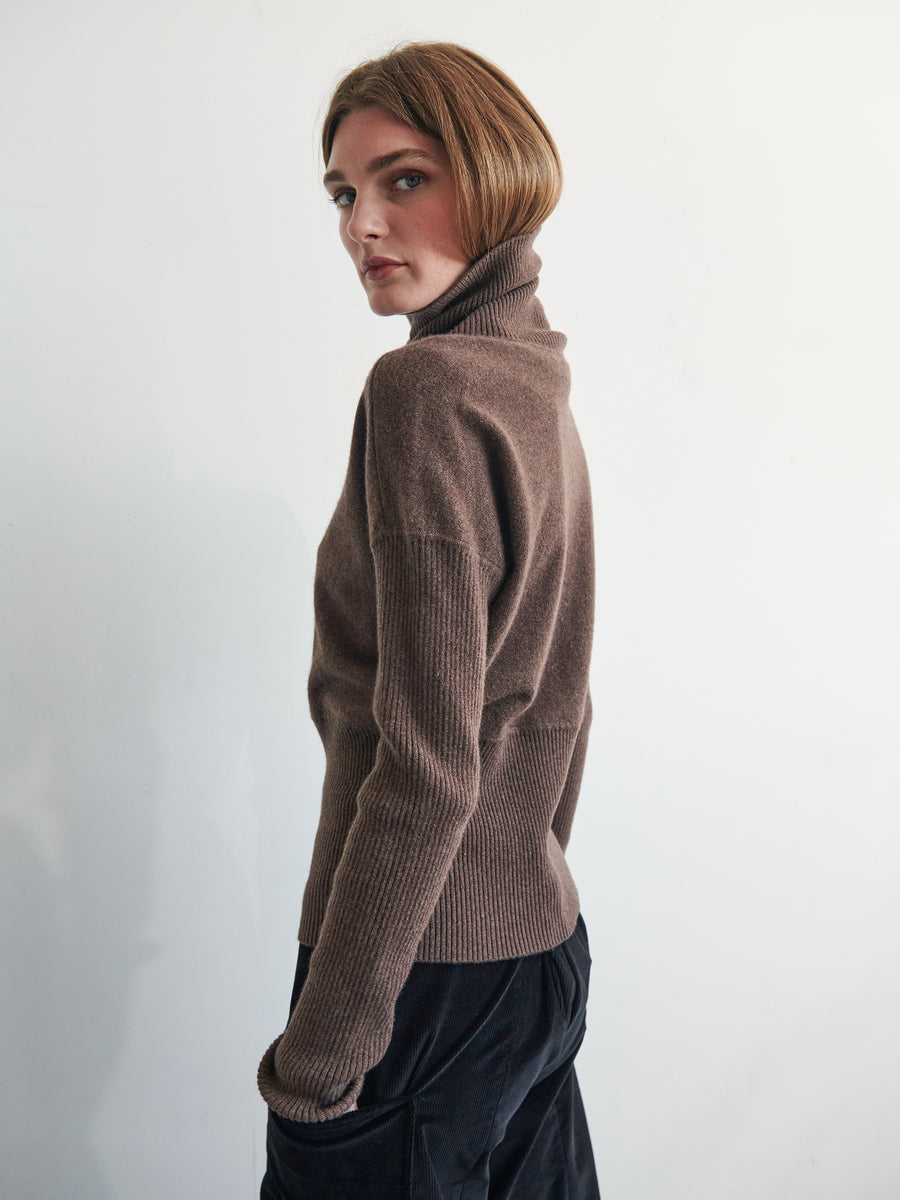 A person with short hair is standing sideways, glancing back over their shoulder. They are wearing a Zero + Maria Cornejo Long-Sleeved Ama Rollneck with ribbed sleeves and black pants. The background is plain and light-colored.