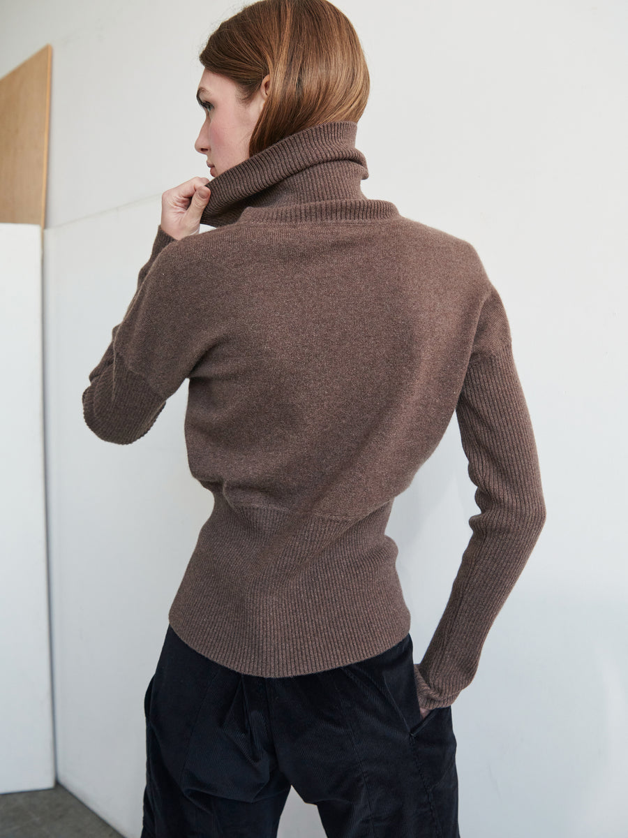 A person with medium-length brown hair is seen from the back. They are wearing a Long-Sleeved Ama Rollneck by Zero + Maria Cornejo and black pants. The background consists of a light-colored wall and a piece of wood leaning against it. The person is holding up the rollneck with one hand, embracing sustainable fashion.