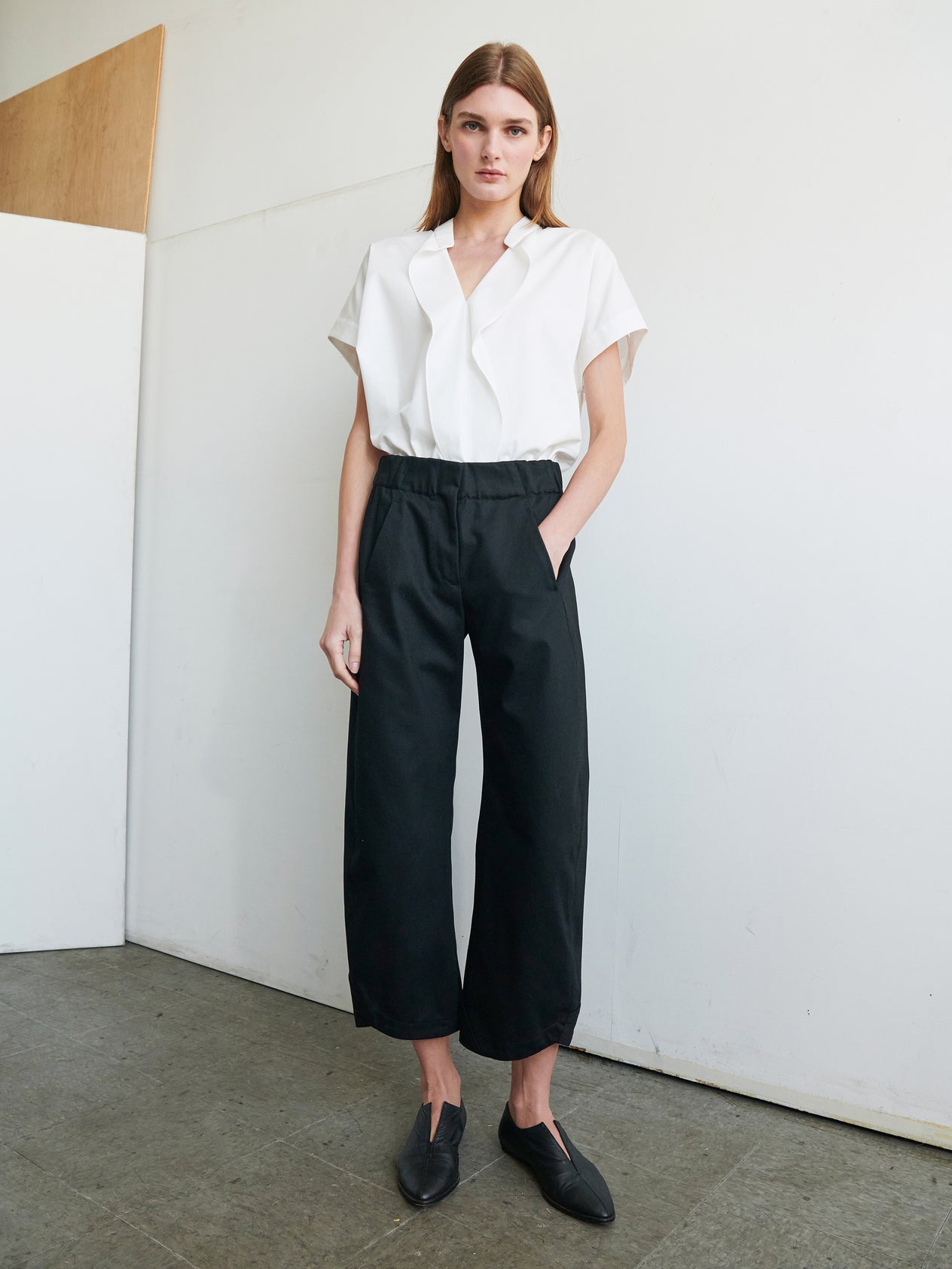 A person with shoulder-length brown hair stands in a relaxed posture against a white wall. They are wearing a white short-sleeved shirt tucked into high-waisted, wide-leg Aissa Cargo Pants by Zero + Maria Cornejo and black slip-on shoes. Their hands are casually in the pockets of their pants.