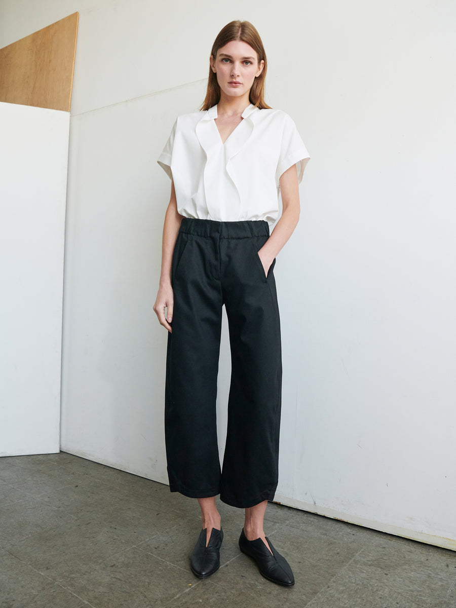 A person with long hair stands against a plain wall, wearing a white short-sleeve blouse tucked into the Aissa Cargo Pant by Zero + Maria Cornejo made from Cradle 2 Cradle Certified™ Gold fabric, paired with black flat shoes. Their hands are in their pockets, and they have a neutral expression.