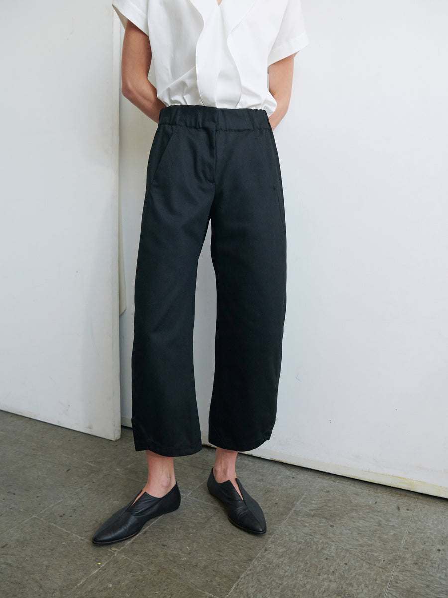 A person is standing against a plain white wall and grey floor, wearing a white short-sleeved shirt, loose-fitting black Aissa Cargo Pants by Zero + Maria Cornejo, and black slip-on shoes. Their arms are crossed and their head is out of frame.