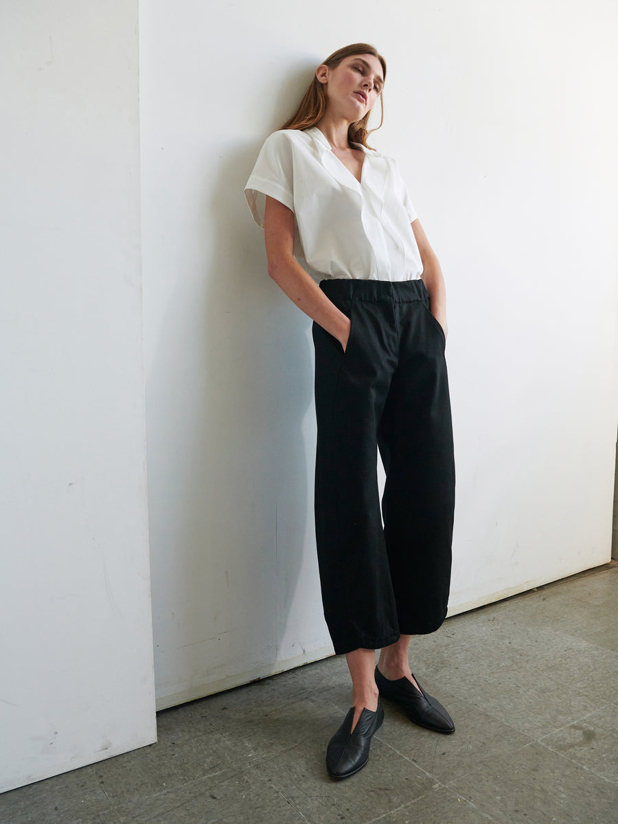 A person stands against a white wall, wearing a loose white blouse tucked into high-waisted Aissa Cargo Pants by Zero + Maria Cornejo. Their hands are in their pockets and they are looking slightly upward. They are also wearing black shoes and their hair is long and straight.