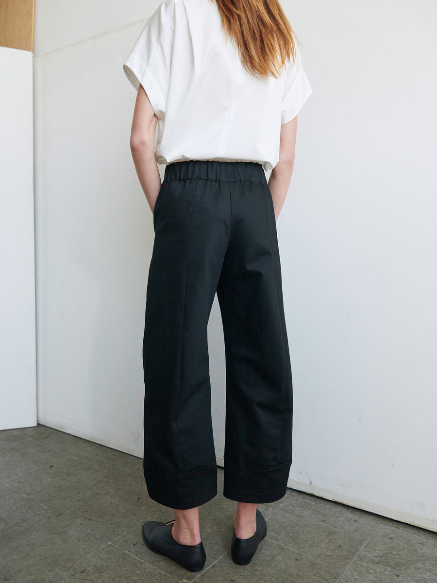 A person with long hair wearing a white short-sleeve shirt and the Aissa Cargo Pant by Zero + Maria Cornejo, stands facing away from the camera. Their hands are in their pockets. They are standing on a gray concrete floor against a plain white background.