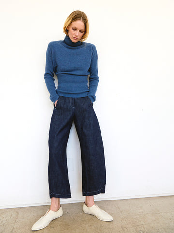 Person with short blonde hair stands against a plain white wall, wearing a blue turtleneck sweater, Aissa Cargo Pants by Zero + Maria Cornejo made from dark organic denim, and light-colored slip-on shoes. They have their hands in their pockets and are looking down.