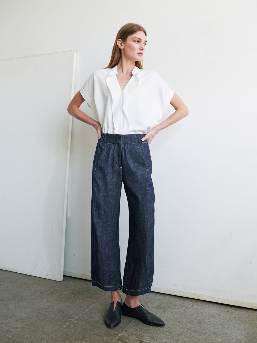 A person with long hair stands against a plain white wall, wearing a white short-sleeved blouse tucked into high-waisted, wide-legged Aissa Cargo Pants from Zero + Maria Cornejo. They are also wearing black loafers. The setting appears to be minimalist and indoors.