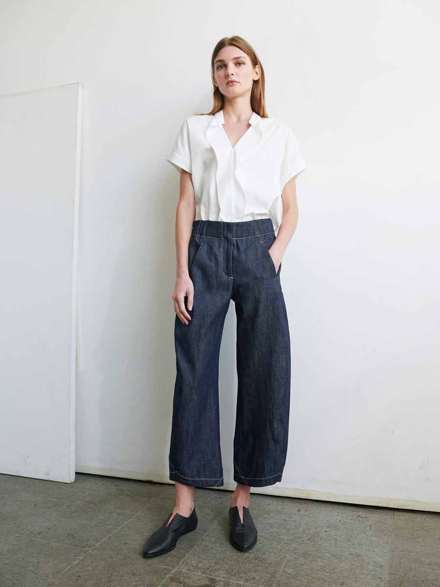 A woman stands against a plain white wall, wearing a white short-sleeve, collared shirt tucked into Zero + Maria Cornejo's high-waisted, wide-leg Aissa Cargo Pant made from organic denim. She has her hands in her pockets and is wearing dark slip-on shoes. The floor is a concrete, industrial-style surface.